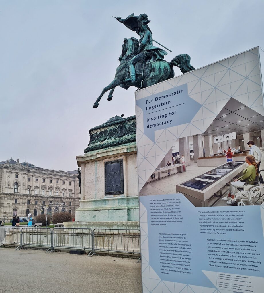 Ein großes Reiterstandbild aus Metall auf einem hohen viereckigen Podest. Es wird teilweise verdeckt durch ein Schaubild, auf dem oben "Für Demokratie begeistern" steht. Darunter ein Foto, das einen modernen, hellen Raum mit Schaukästen, Tischen und mehreren jungen Menschen, eine Frau davon einem Rollstuhl, zeigt. Darunter Text. Links vom Denkmal ist ein Teil der Hofburg zu sehen.