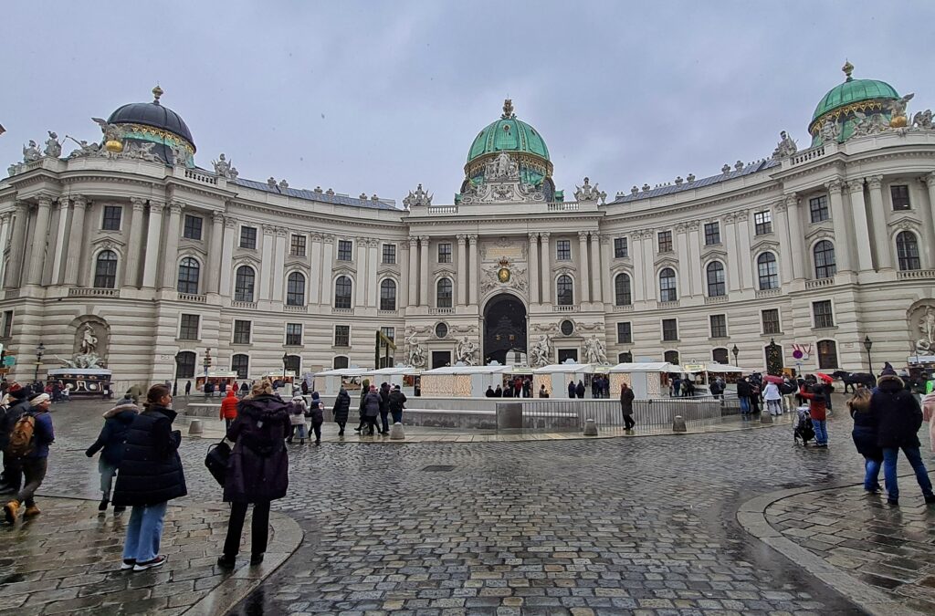 Ein prächtiges helles Gebäude mit mehreren Stockwerken und gleichmäßig angeordneten Fenstern. Es erstreckt sich nach beiden Seiten hinter einem großen Platz, auf dem viele Menschen gehen oder stehen. Direkt vor dem Gebäude stehen außerdem mehrere Weihnachtsmarktstände. Über dem hohen Eingangstor in der Mitte und an beiden Seiten sind oben kleine, breite Türme mit grünen Kuppeln. Davor sind auf dem Dachbereich viele Figuren und Statuen.