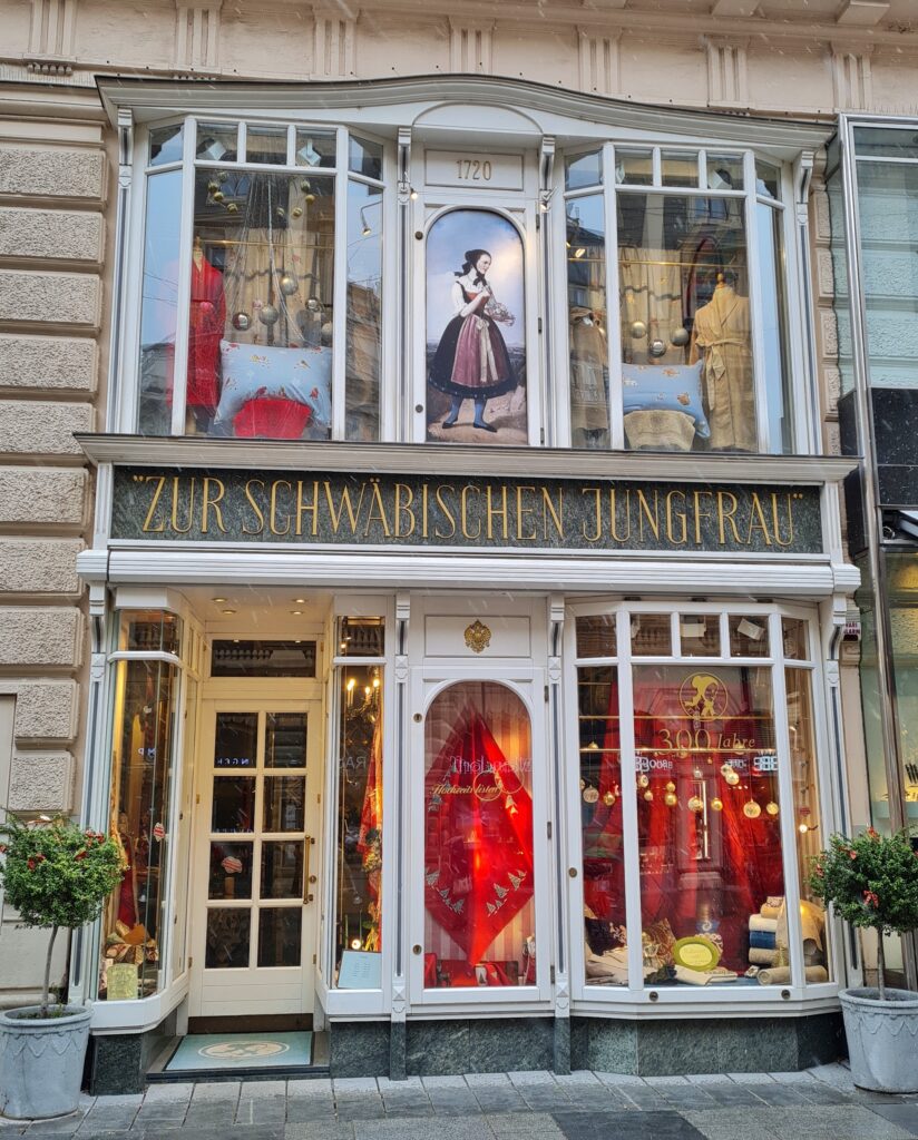 Der untere Teil eines Gebäudes mit einer hohen, schmalen Eingangstür und einem kleinen Schaufenster daneben. Oben weitere schmale Schaufensterflächen, und in der Mitte ein Gemälde einer jungen Frau in Tracht. Darüber steht "1720". Zwischen den den Stockwerken in altmodischer Schrift "Zur schwäbischen Jungfrau"