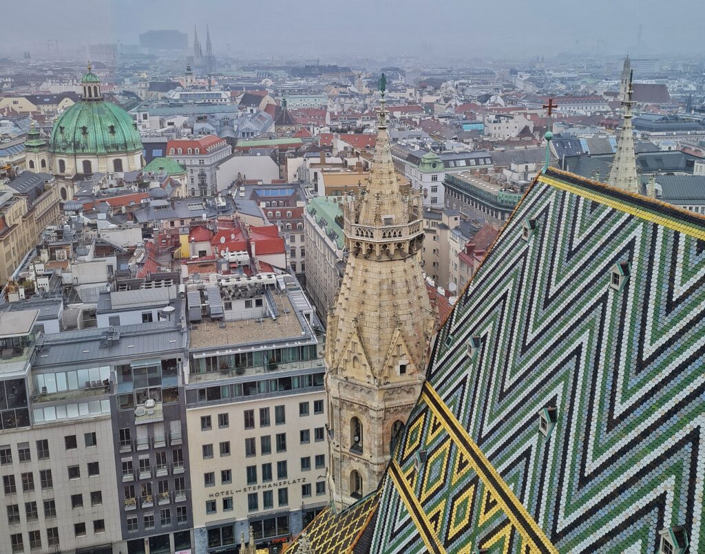 Blick über die Stadt mit hohen Häusern und Straßenschluchten. Im Hintergrund eine Kirche mit zwei Türmen, links eine grüne Kuppel. Im Vordergrund sieht man einen Teil des bunten Daches des Stephansdomes mit Zickzack-Muster und einen kleinen Turm an der Ecke.