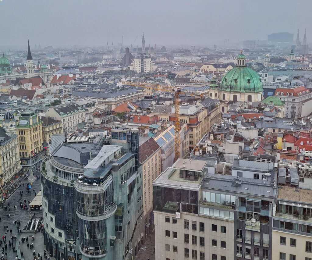 Blick auf die Stadt von oben. Im Vordergrund zwei hohe moderne Gebäude, dahinter viele ältere mehrstöckige Häuser und Kirchen. Eine davon ist hell mit einer runden grünen Metallkuppel.