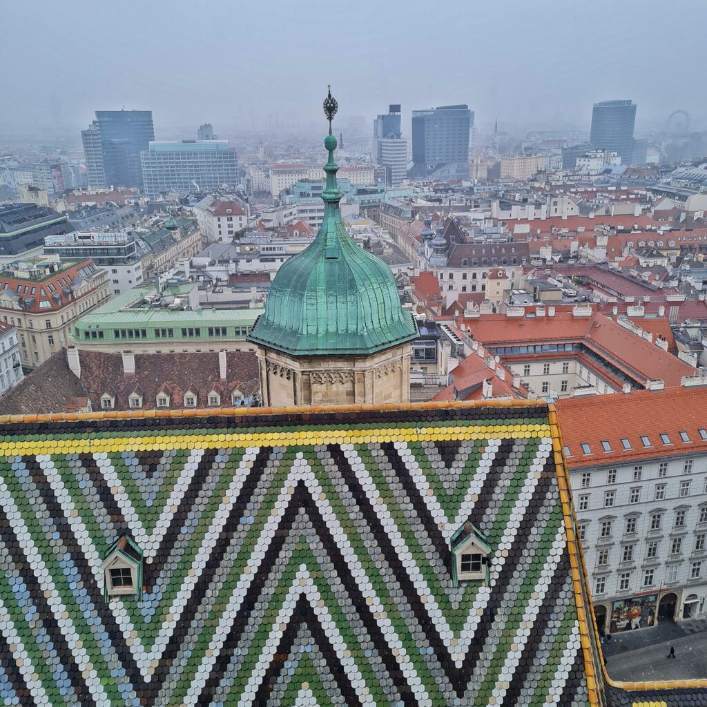 Blick von oben über die Stadt. Vorne ein Teil des bunten Daches des Domes. Dahinter ein Turm mit einer grünen Metallspitze. Rechts davon sieht man ein mehrstöckiges Gebäude, das mit seinen vier Seiten einen Innenhof umfasst. Dahinter weitere Gebäude, ganz im Hintergrund einige moderne Hochhäuser.