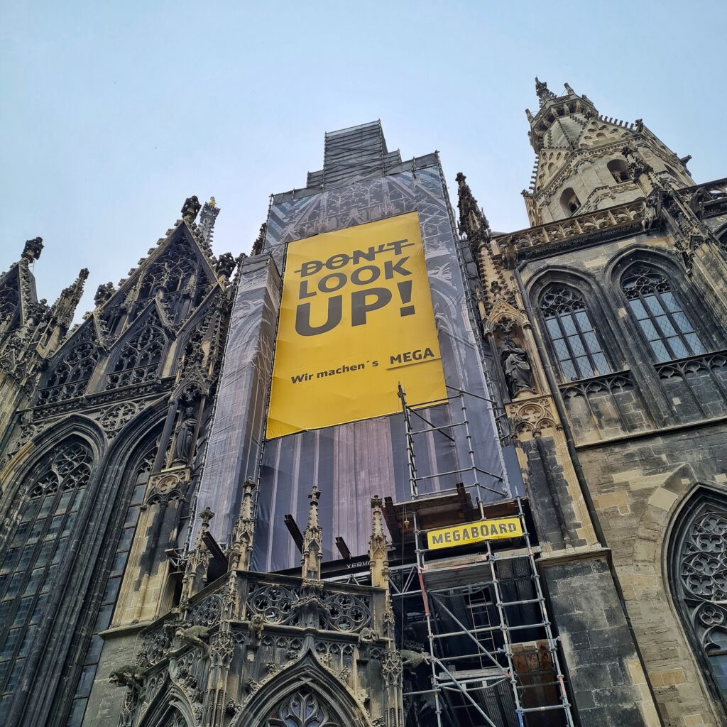 Seitliches Foto vom Stephansdom, von schräg unten fotografiert. Man sieht die verzierte Fassade, hohe Fenster, einen Turm und ein Gerüst, an dem ein großes gelbes Transparent hängt. Auf diesem steht durchgestrichen "Don't" und darunter "Look Up!"; ganz unten steht "Wir machen's Mega".