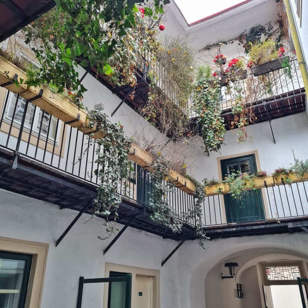 Blick in den Innenhof eines weißen Gebäudes. Unten sind die oberen Teile von Haustüren und einem Torbogen zu sehen. Darüber in zwei Stockwerken durchgehende Balkone vor dunkelgrünen Wohnungstüren. Über die Geländer hängen grüne Pflanzen darunter. An den oberen Pflanzen hängen rote Weihnachtskugeln und künstliche rote Blumen.