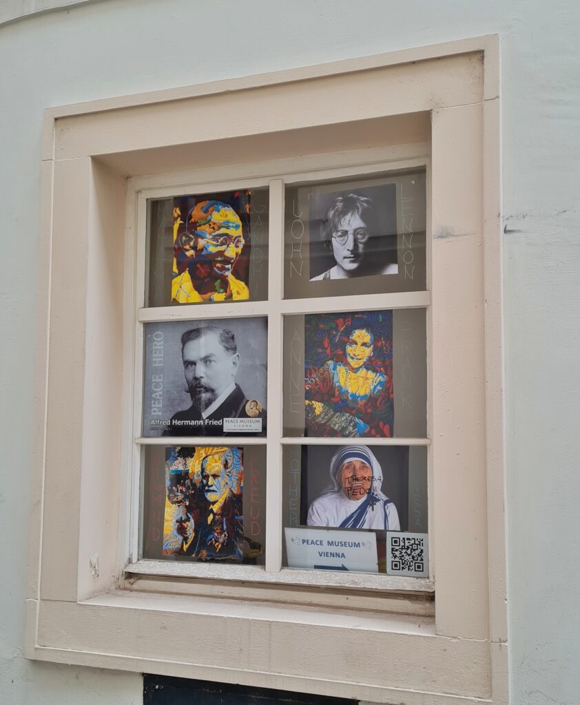 Ein Sprossenfenster mit beigem Rahmen. Die sechs kleinen Fensterflächen sind von Porträtfotos verdeckt, zum Beispiel von Mahatma Ghandi, John Lennon und Mutter Theresa. An der unteren linken Fensterfläche hängt ein kleines Schild : Peace Museum Vienna.