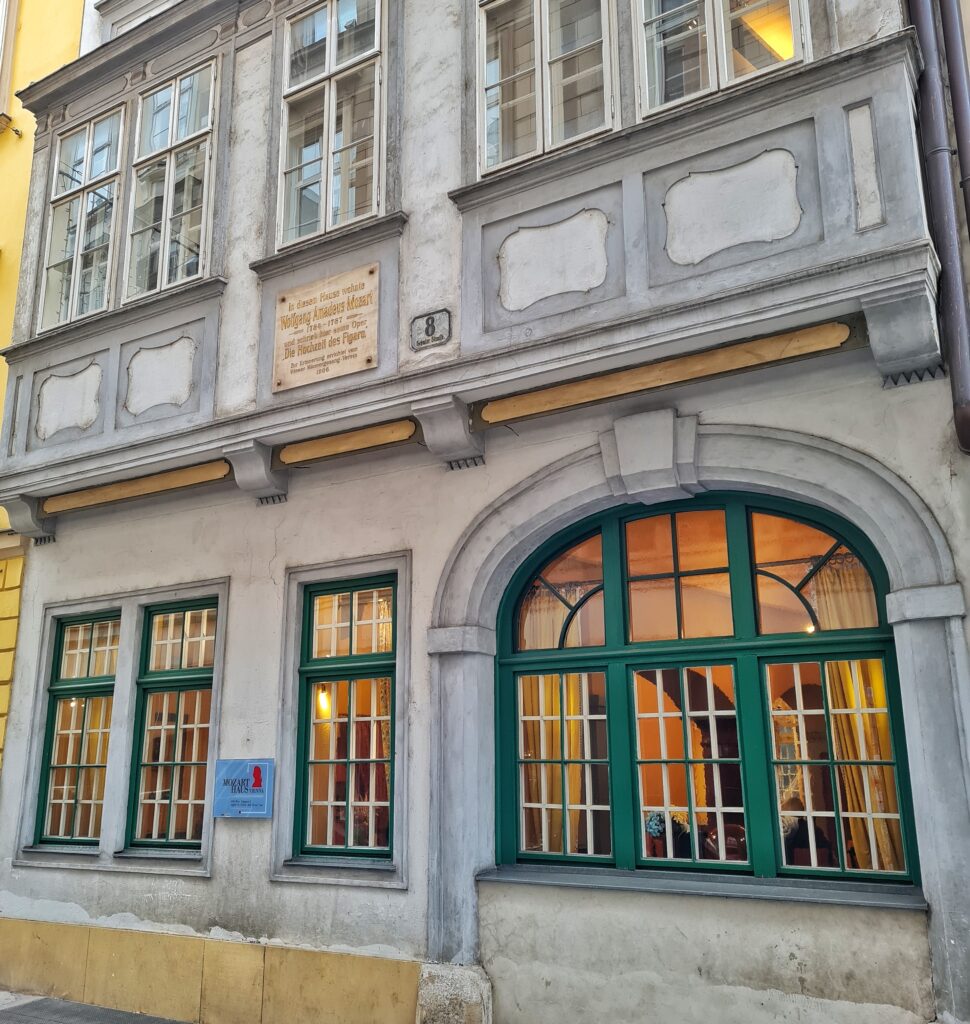 Ein gräulich-weißes Gebäude. Unten sind Sprossenfenster und ein größeres halbrundes Fenster mit dunkelgrünen Rahmen. Darüber weiße Vierecke auf grauem Hintergrund und in der Mitte ein Schild: "In diesem Haus wohnte Wolfgang Amadeus Mozart und schrieb hier seine Oper Die Hochzeit des Figaro". Im oberen Geschoß weitere Sprossenfenster mit weißlichen Rahmen.