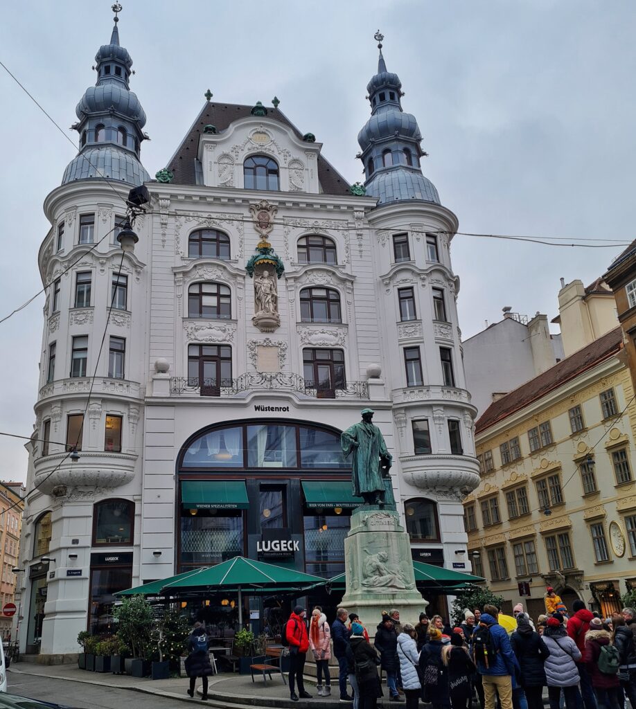 Ein mehrstöckiges hohes Gebäude. An den beiden Seiten sind runde Anbauten, die auf dem Dach in Metalltürmchen übergehen. Unten ist eine größere Fensterfassade, auf der "Lugeck" steht. Davor große, dunkelgrüne Sonnenschirme, ein Denkmal und eine Gruppe von Menschen.