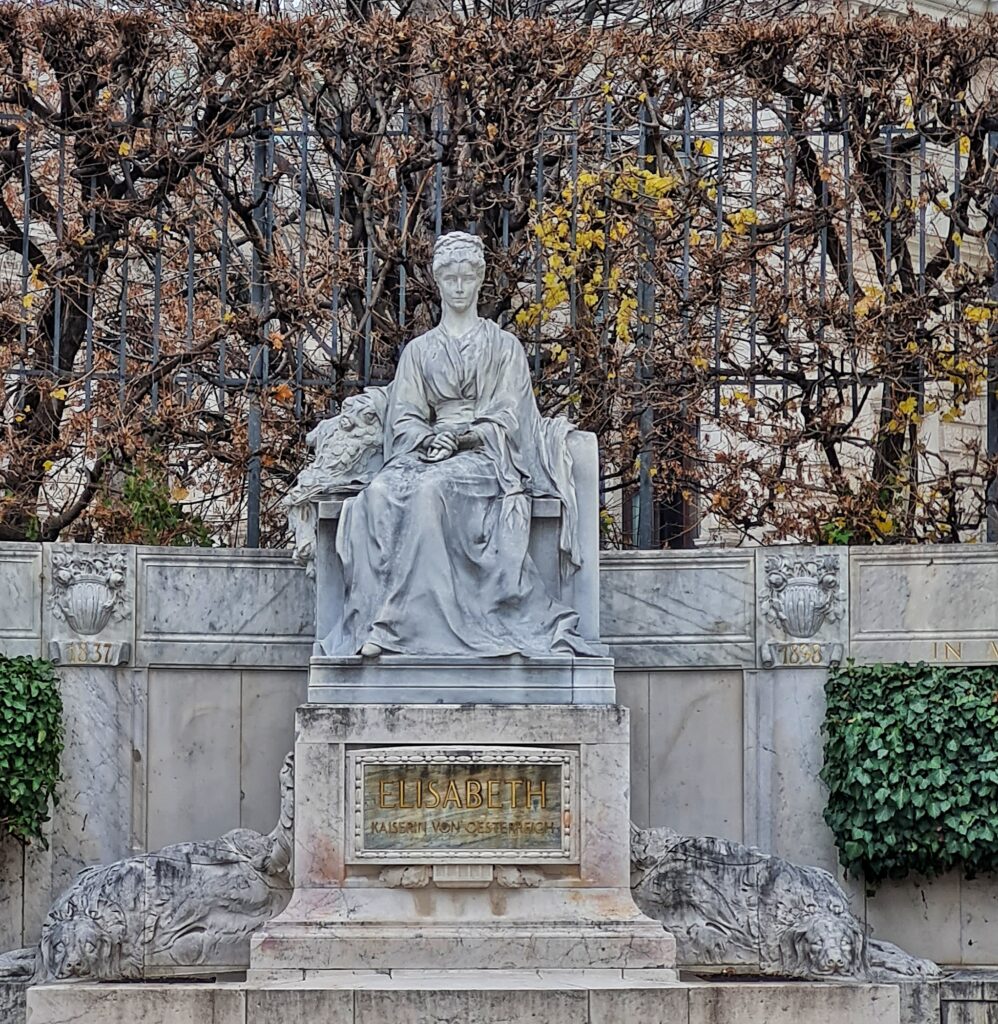 Vor einer Steinmauer und spärlich belaubten Bäumen das steinerne Denkmal einer sitzenden Frau in wallenden Gewändern. Auf dem Podest darunter steht: "Elisabeth, Kaiserin von Österreich".