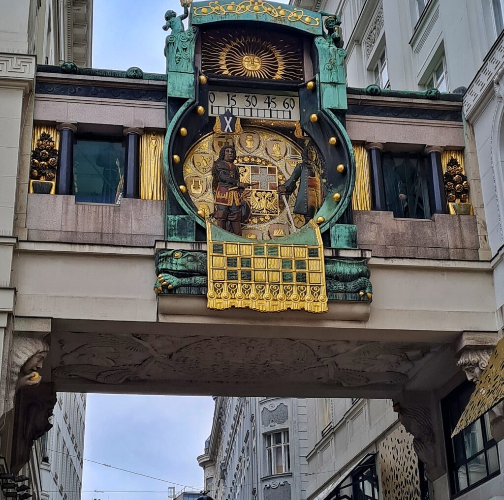 Ein geschlossener Durchgang quer über eine schmae Gasse. Daran hängt eine kunstvoll geschmückte Uhr aus grünem Metall mit Gold. Zentral in der Mitte ist ein Kreis mit Symbolen statt der Ziffern und kunstvollen Figuren, Darüber eine Linie mit den Zahlen 0, 15, 30, 45 und 60. Ein schwarzes X steht kurz vor der 15.