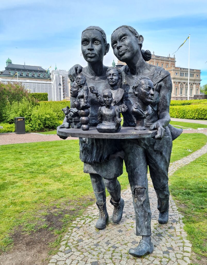 Zwei Bronzefiguren, die in Gehbewegung zur Kamera hin dargestellt sind. Es handelt sich um zwei Frauen mit groben Gesichtszügen und einfacher, teilweise kaputter Kleidung. Sie tragen gemeinsam eine flache Platte, auf der Büsten von Frauen stehen. Sie stehen auf einem gepflasterten Weg in einem kleinen Park mit Rasenflächen und niedrigen Büschen.
