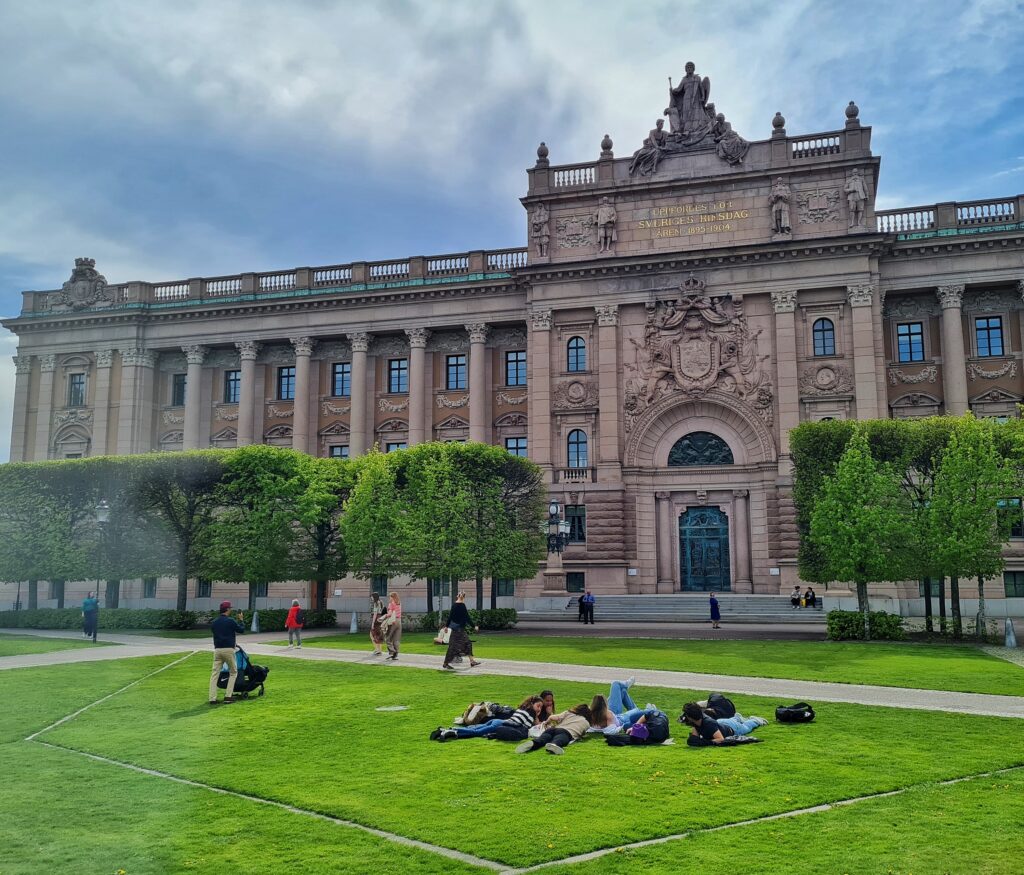 Das Bild zeigt ein großes, prächtiges Gebäude mit einem grünen Rasen davor. Das Gebäude ist aus Stein und hat viele Fenster und Säulen. Der Eingang des Gebäudes ist groß und prächtig und hat eine Statue oben drauf. Der Rasen ist ordentlich gepflegt und hat ein paar Bäume. Es gibt ein paar Menschen auf dem Rasen, einige sitzen und einige liegen; andere laufen auf den Wegen dazwischen. Der Himmel ist blau mit ein paar Wolken.