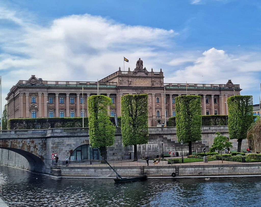 Das Bild zeigt ein großes, prächtiges Gebäude aus hellbraunem Stein mit vielen Säulen und Statuen an der Fassade. Das Gebäude ist von Bäumen und Grün umgeben und befindet sich am Ufer eines Flusses. Der Fluss im Vordergrund hat eine Steinbrücke und ein kleines Boot. Der Himmel ist blau mit weißen Wolken. Das Bild scheint aus der Ferne aufgenommen worden zu sein und gibt einen weiten Blick auf das Gebäude und seine Umgebung.Das Bild zeigt ein großes, prächtiges Gebäude aus hellbraunem Stein mit vielen Säulen und Statuen an der Fassade. Das Gebäude ist von Bäumen und Grün umgeben und befindet sich am Ufer eines Flusses. Der Fluss im Vordergrund hat eine Steinbrücke und ein kleines Boot. Der Himmel ist blau mit weißen Wolken.
