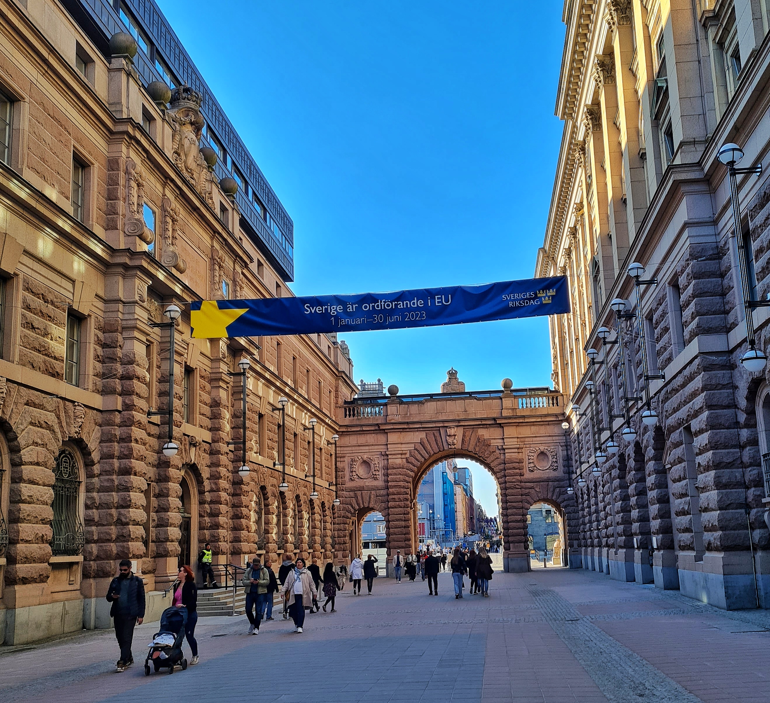 Ein breiter Durchgang mit alten, prächtigen Gebäuden auf beiden Seiten und einem großen Torbogen im Hintergrund. Mehrere Spaziergänger laufen zwischen den Gebäuden hindurch. Darüber hängt ein blaues Banner mit der Aufschrift “Sverige är ordförande i EU” (Schweden ist der Vorsitzende der EU). Der Banner hat einen gelben Stern darauf. Der Himmel ist blau und das Wetter ist klar.