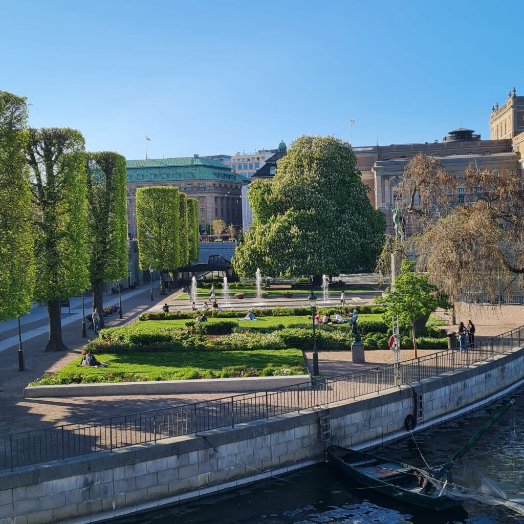 Ein Park mit Rasenflächen, Blumen, niedrigen Büschen, Bäumen, einem Springbrunnen mit mehreren kleinen Fontänen und einer Statue. Eine Reihe von Bäumen auf der rechten Seite sind viereckig beschnitten. Mehrere Menschen laufen auf den Wegen oder sitzen auf dem Rasen. Im Hintergrund sind große, alte Gebäude zu sehen. Rechts vorne ist eine Wasserfläche mit einer Kaimauer zu sehen. Dort ist ein Ruderboot angebunden.