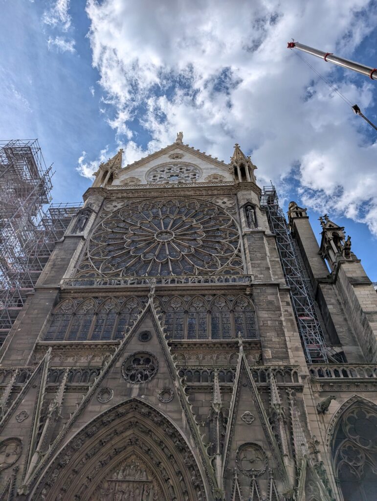 Blick auf die gotische Fassade der Kathedrale Notre-Dame de Paris, teilweise eingerüstet, mit Wolken am blauen Himmel.